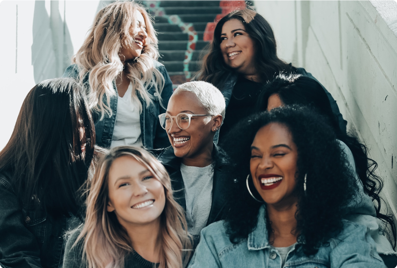 A group of women of diverse ethnic backgrounds sitting together on a staircase, smiling at one another.