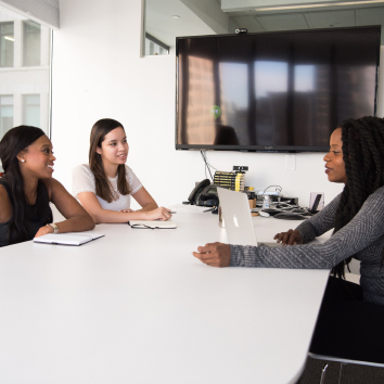 Three women sitting at a conference table, smiling. One is looking at a laptop screen.