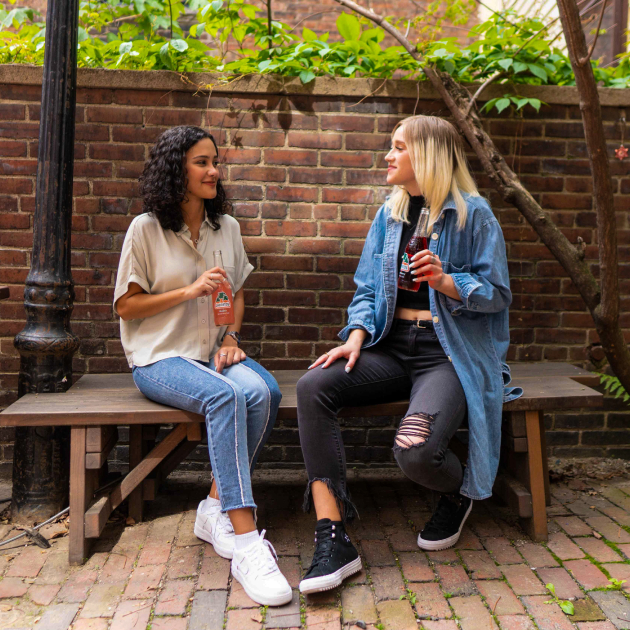 Two women sitting on a bench in front of a brick wall looking and smiling at each other, each holding a glass soda bottle.