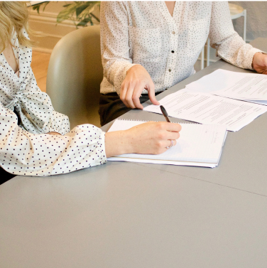 Two people sitting at a desk with papers in front of them. One person is writing something on the paper.