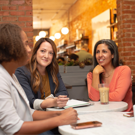 Three women sitting together at a table inside of a cafe. They are smiling.